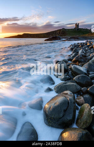 Les ruines de château de Dunstanburgh perchées sur la falaise au-dessus de Embleton Bay comme la mer du Nord autour de la pause vagues rochers dolérite le long du rivage. Banque D'Images