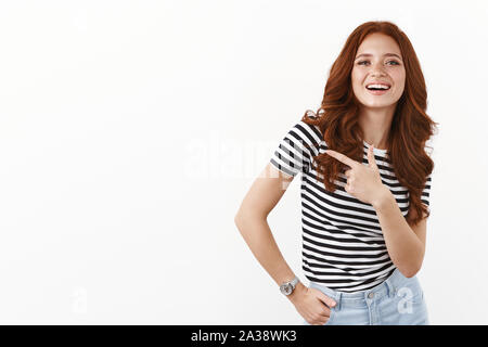 Jeune étudiante moderne sans souci en t-shirt à rayures avec du gingembre curly hairstyle, sourire sûr de lui, montre de l'index gauche, aider à trouver l'espace de copie Banque D'Images