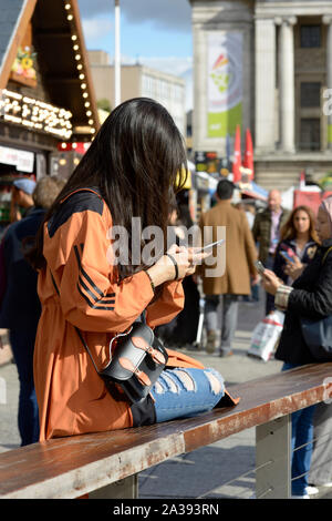 Orientale, en manteau beige et jeans déchirés, ce qui rend vos autoportraits, assis sur un banc Banque D'Images