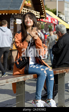 Orientale, en manteau beige et jeans déchirés, ce qui rend vos autoportraits, assis sur un banc Banque D'Images