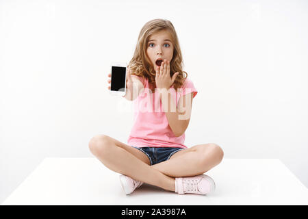 Cute blonde jeune offres choqué teenage girl sitting jambes croisées sur le plancher, bouche ouverte haletant inquiets, regarder l'écran du smartphone, montrer l'appareil photo partager Banque D'Images