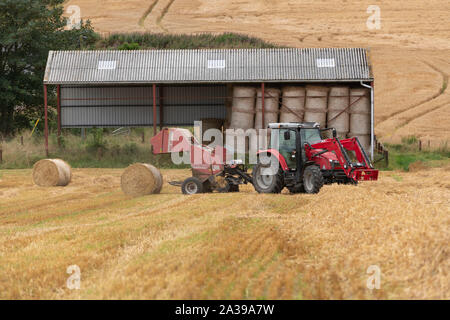 Un tracteur et la ramasseuse-presse-une botte de paille sur une colline en face d'un Dutch Barn contenant plus de bottes de paille Banque D'Images