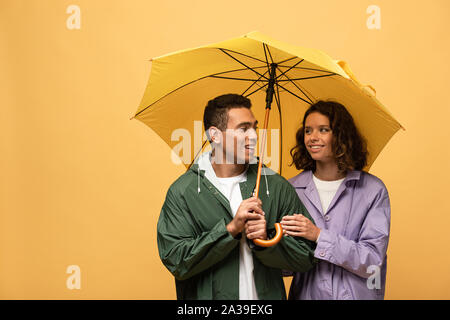 Smiling couple interracial dans d'imperméables holding umbrella isolé sur le jaune Banque D'Images