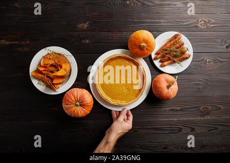 Vue partielle de l'homme main à côté de la plaque avec la citrouille, cuite ensemble carotte, coupée en rondelles et l'ensemble de la table en bois sombre sur les citrouilles Banque D'Images