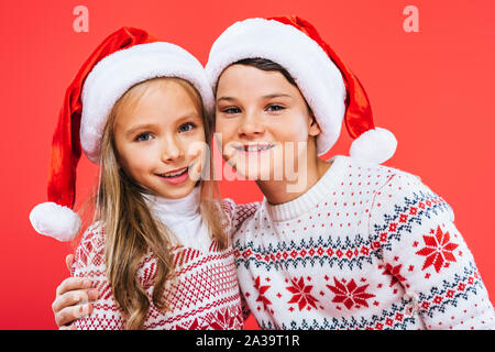 Vue de face de deux enfants souriant à santa hats et les chandails embrassant isolé sur red Banque D'Images