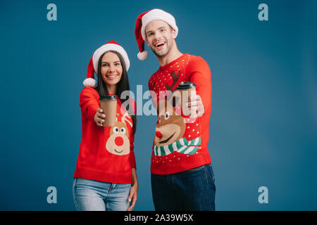 Cheerful couple dans santa hats et chandails de noël holding coffee pour aller isolé sur blue Banque D'Images