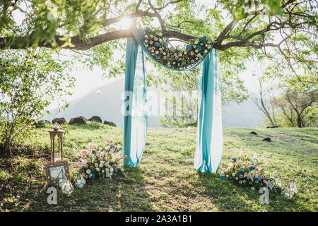 Cérémonie de mariage Sunset arch avec décoration de fleurs et tissu bleu accroché sur la canopée grand arbre. Hortensia bleu, rose, Lily. De couleur pastel. Banque D'Images