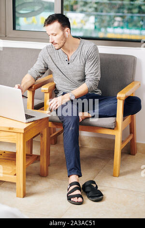 Beau jeune homme portant des écouteurs sans fil lors de la vérification des e-mails sur l'ordinateur portable Banque D'Images