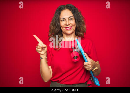 L'âge moyen du patineur senior woman holding skateboard sur fond isolé rouge très heureux pointant avec la main et le doigt sur le côté Banque D'Images