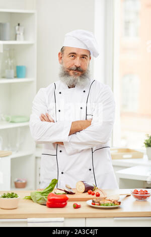 Taille portrait de cheerful senior chef looking at camera standing with arms crossed in restaurant kitchen Banque D'Images