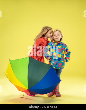 Un portrait d'un lumineux à la mode des filles dans un imperméable tenant un parapluie arc-en-ciel de couleurs sur fond jaune studio. L'automne et au printemps, à la mode pour enfants. Cute blonde élégant. Banque D'Images