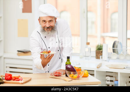 Portrait of cheerful senior chef presenting beau bol en posant dans une cuisine de restaurant, copy space Banque D'Images