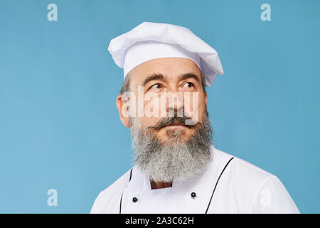 Head and shoulders portrait de barbu charismatique chef looking up permanent tout en se posant contre fond bleu, copy space Banque D'Images