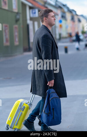 Homme marchant le long d'une rue avec ses bagages transportant un sac et en tirant une valise derrière lui dans un concept de voyage Banque D'Images