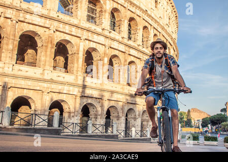 Heureux jeune homme avec le vélo touristique portant chemise et hat au Colisée à Rome, Italie au lever du soleil. Banque D'Images