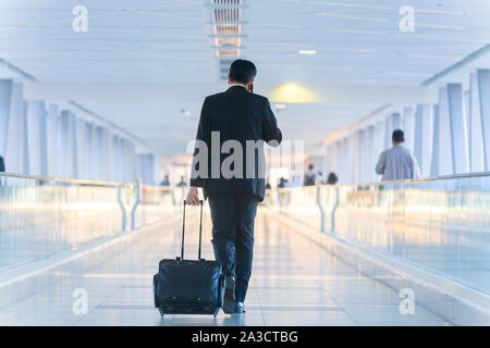 Businessman walking et roulant un chariot valise dans le hall, parlant sur un téléphone mobile. Concept de voyages d'affaires. Banque D'Images