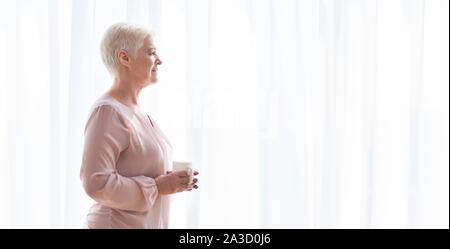 Pensive senior lady avec mug d'un séjour près de window Banque D'Images