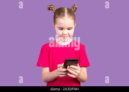 Petite fille, dans un lumineux T-shirt, utilise un smartphone isolé sur fond violet. Banque D'Images