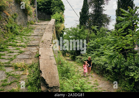 Carefree woman and girl holding hands et flâner le long chemin herbeux entouré par de vieux parapet avec des arbres verts Banque D'Images