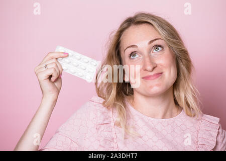 Portrait of young blonde woman wearing pink dress holding blister de médicament comprimés à up smiling. Studio shot sur fond rose Banque D'Images