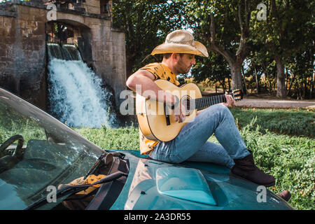 L'âge moyen homme portant un chapeau de cowboy assis sur un véhicule tout-terrain tout en jouant de la guitare à l'extérieur Banque D'Images