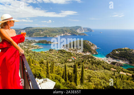 Femme en robe rouge longue portant un chapeau tenant un cocktail debout sur un balcon au-dessus de la Bella Vista Resort grecque de Paleokastritsa Corfu Grèce Banque D'Images