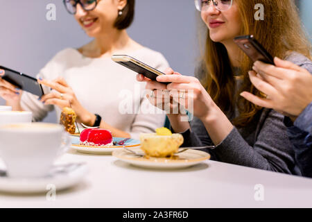 La technologie, mode de vie, l'amitié et de personnes concept - trois jeunes femmes heureux avec les smartphones de faire des photos de leurs tasses à café et desserts au Banque D'Images