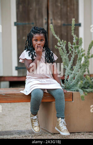 Jeune fille noire avec les doigts dans sa bouche assis sur banc en bois Banque D'Images