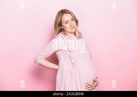 Jeune femme enceinte portant robe rose debout tenant son ventre tout en gardant une main sur son dos. Studio shot sur fond rose with copy space Banque D'Images