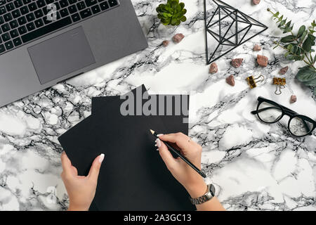 Main d'une femme avec une manucure, bague et montre est la tenue de deux feuilles de papier noir et d'un crayon, allant d'écrire quelque chose. Ordinateur portable, lunettes, classique Banque D'Images