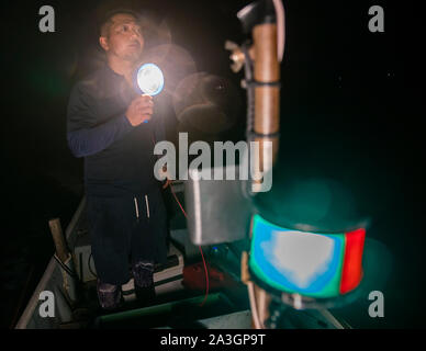 Pêche à la ligne avec harpon Takiya-ryo. Le pêcheur Ikuya Yamamoto tient une lampe portable brillante et regarde vers le haut la nuit sur le lac Hamana près de Hamamatsu, au Japon, la lumière de navigation verte du bateau floue en arrière-plan. Banque D'Images
