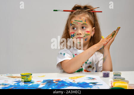 Petite fille en t-shirt blanc, avec des pinceaux dans ses cheveux sont assis à la table avec les peintures et Whatman, peinture sur elle. Isolé sur blanc. Close-up. Banque D'Images