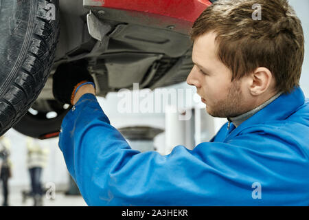 Homme aux cheveux brun avec la lumière de l'examen de l'unité de suspension de la voiture automobile levé . Expert compétent en uniforme bleu et gants noirs fixant les pièces cassées de la voiture à la place de travail Banque D'Images