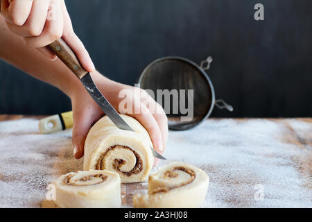 Woman's hands cutting cannelle maison rouler la pâte sur une surface enfarinée avec duster en arrière-plan. Focus sélectif avec avant-plan flou Banque D'Images