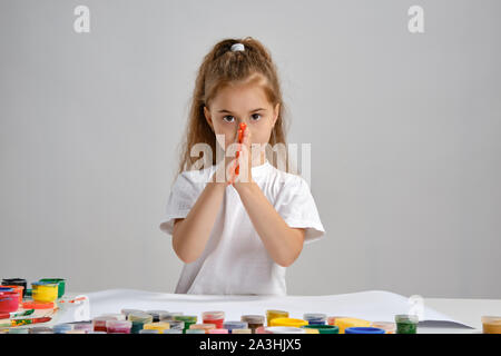 Petite fille en t-shirt blanc assis à table avec des peintures colorées et Whatman, peindre ses mains. Isolé sur blanc. Medium close-up. Banque D'Images