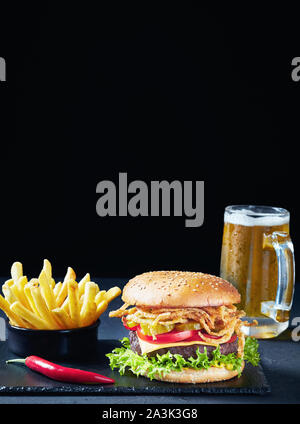 Close-up of Cheeseburger avec fromage cheddar, oignons frits croustillants, laitue, tomates, cornichons, frites et de la bière dans un verre une tasse dans le bla Banque D'Images