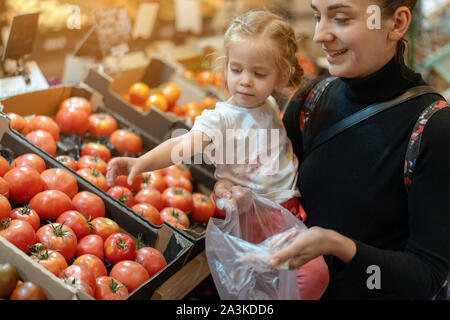 Happy smiling young woman with little daughter acheter tomates rouges sur le marché. La mère et l'enfant le choix de légumes de saison en supermarché. Fam Banque D'Images