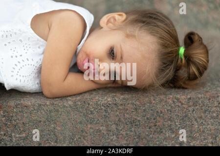 Une belle petite fille de l'apparition de cheveux blond couché sur l'asphalte de la rue triste. Les problèmes des enfants.Enfant fatigué des caprices. Banque D'Images