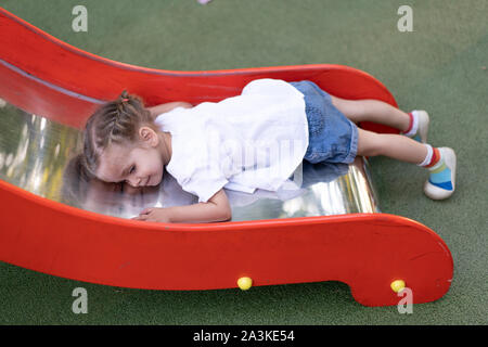 Peu Caucasian girl se trouve sur une diapositive à l'aire fatigué d'une triste journée d'été. Les enfants fatigués tendre enfance. de loisirs d'été. Banque D'Images