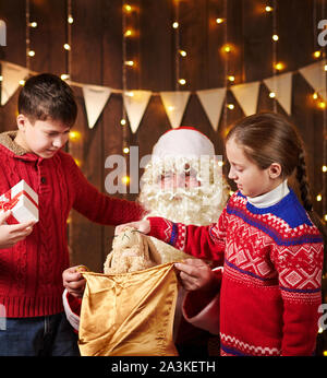 Le Père Noël ouvre un sac et donne des cadeaux aux enfants, assis près de l'arbre de Noël décoré avec des lumières - Joyeux Noël et bonnes fêtes ! Banque D'Images