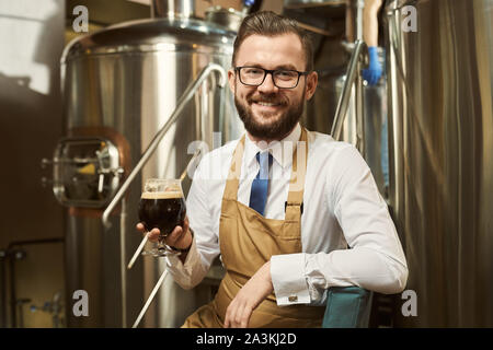 Positif, cheerful brewer on metal plate-forme, tenant le verre à bière brune. Beau worker smiling et posant sur fond de réservoir de stockage en acier. Banque D'Images