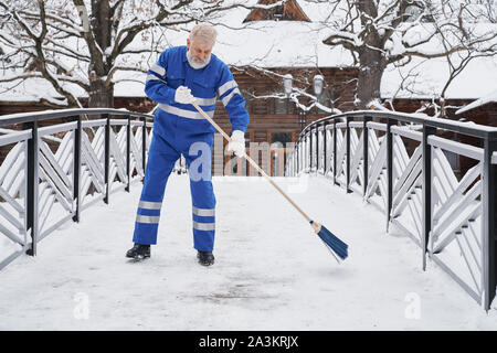 Vue de face de l'homme graves portant des uniformes bleu debout sur pont et enlever la neige en hiver. Homme barbu, balai de maintien et de nettoyage de sentier dans la matinée. Concept de compensation. Banque D'Images