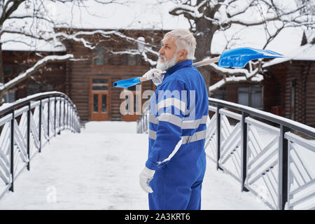 Voir du côté de l'homme barbu portant des uniformes bleu pelle maintien sur l'épaule et à la de côté en hiver. Travailleur masculin en processus de nettoyage pont et sentier de neige. Concept de service. Banque D'Images