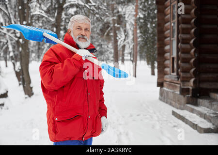 Beau grand-père wearing red jacket pelle maintien dans l'épaule, looking at camera and smiling in hiver froid. Homme debout en position de stationnement et de nettoyage de sentier de neige. Concept de compensation. Banque D'Images