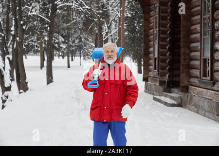 Vue avant du grand-père barbu wearing red jacket looking at camera et souriant tout en gardant l'épaule sur la pelle en hiver. L'homme travaillant dans les rues du parc et le nettoyage de la neige. Concept de service. Banque D'Images