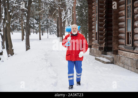 Homme barbu wearing red jacket holding pelle, looking at camera et aillent de l'avant en position de stationnement. Edler homme et le nettoyage des rues de la neige et de la glace. Concept de propreté et l'emploi. Banque D'Images