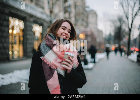 Happy young woman with disposable cup portant des vêtements chauds en marchant sur la rue de la ville en hiver Banque D'Images