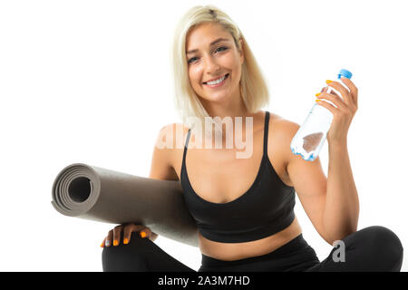 Une jeune fille sportive avec des cheveux blonds et lumineux dans une manucure sports top noir et leggings détient un tapis de sport et une bouteille d'eau. Banque D'Images