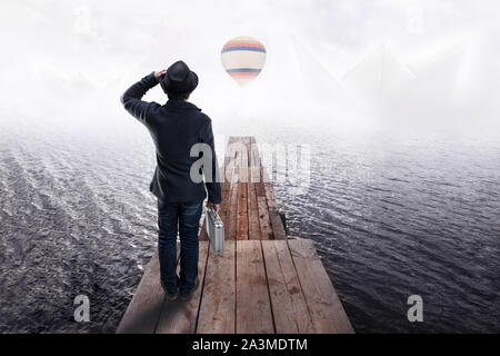 Vue arrière d'un homme en chapeau noir, debout sur la jetée en bois et à la recherche dans la distance à partir de bateaux en papier et colorés en montgolfière. Concept de rêves et Banque D'Images