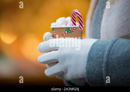 Close-up woman's hands holding gants de tasse de chocolat chaud avec de la guimauve. Contexte de l'éclairage d'enseignes lumineuses et décorées juste des kiosques et shop Banque D'Images